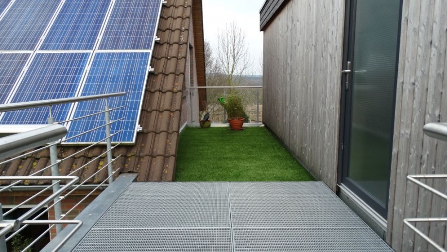 Moderne Dachterrasse mit Solarmodulen, Kunstrasen, Blumentöpfen und einer Holzwand.