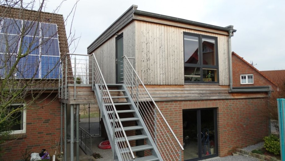 Modernes zweistöckiges Gebäude aus Ziegel und Holz mit Solarpaneelen und Metalltreppe, bewölkter Himmel.