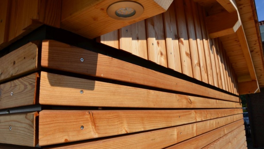 Holzfassade mit warmen Brauntönen, sichtbarer Maserung und einer kleinen Lampe unter einem Vorsprung.
