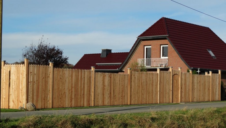 Ein Backsteinhaus mit rotem Dach hinter einem hohen Holzzaun, umgeben von grünem Gras und Bäumen.