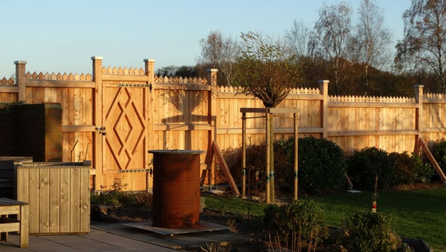 Ein gepflegter Garten mit Holzzaun, rostigem Metallfass, Holzbank und einem neu gepflanzten Baum.