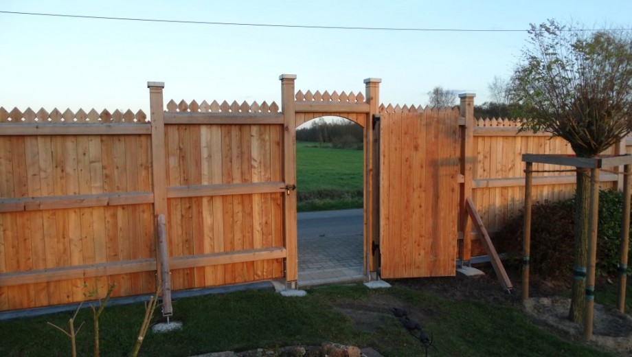 Ein offenes Holztor führt von einem Garten zu einer ruhigen Landstraße, umgeben von grünen Wiesen.