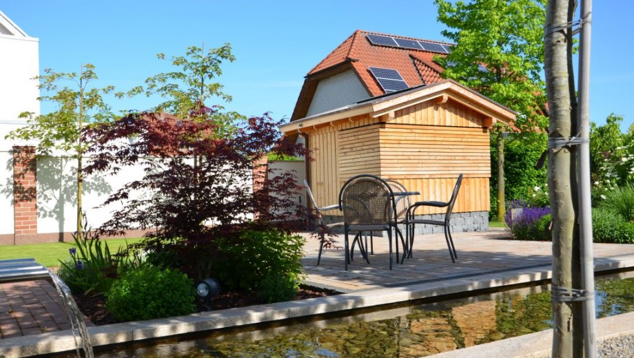 Idyllischer Garten mit Holzhaus, Solarpaneelen, Tisch, Stühlen und kleinem Teich unter blauem Himmel.