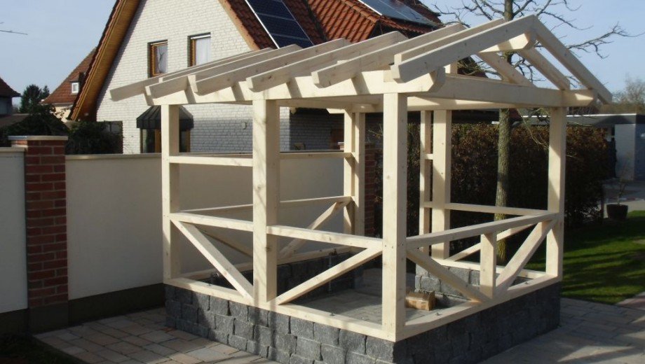 Holzkonstruktion im Bau auf Steinbasis, umgeben von Ziegelmauer und Haus mit Solarpaneelen.