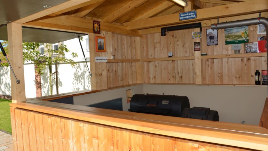 Ein gemütlicher, überdachter Grillplatz aus hellem Holz mit dekorativen Schildern und einem großen Grill.