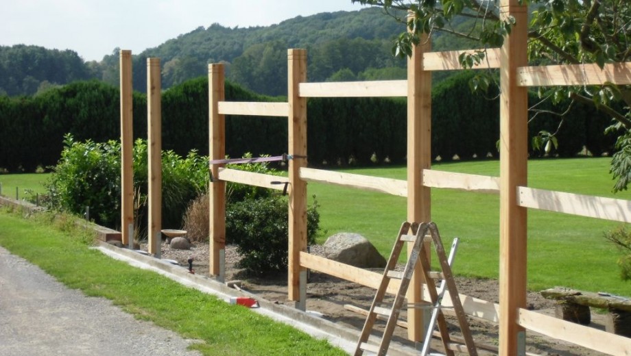 Eine Baustelle zeigt einen unvollständigen Holzzaun mit Werkzeugen, umgeben von grünem Rasen.