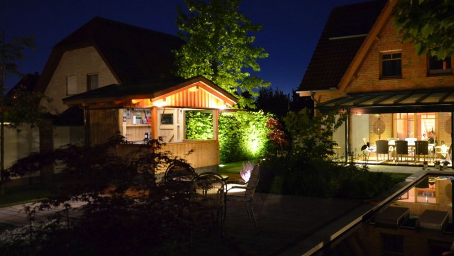 Nächtlicher Garten mit beleuchtetem Pavillon, Terrasse und Haus, umgeben von Pflanzen und Bäumen.