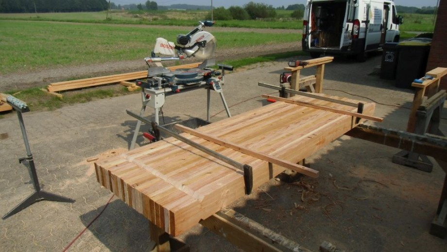 Ein Handwerker arbeitet im Freien an einem Holztisch mit einer Bosch-Gehrungssäge, umgeben von Natur.