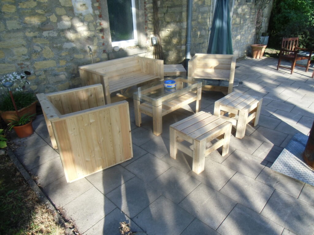 Terrasse mit hellen Holzmöbeln, darunter Sitzbank, Tisch und Hocker, umgeben von Pflanzen und Steinmauer.