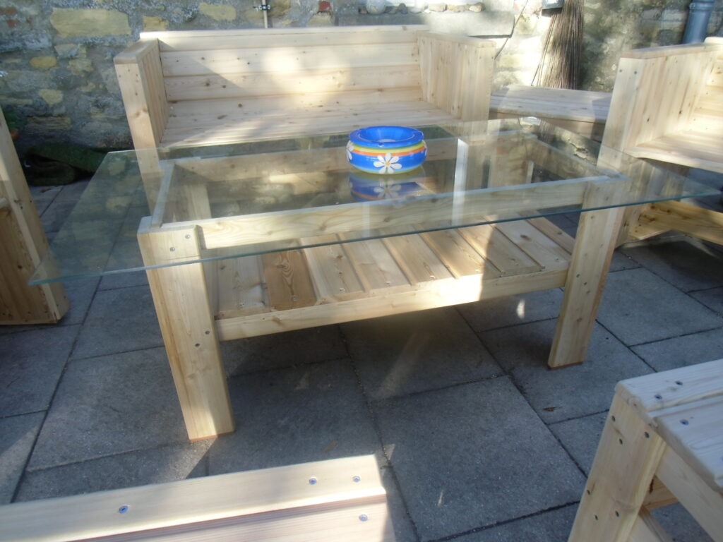 Rustikale Holz-Sitzgruppe mit Glastisch und bunter Schale, umgeben von grauen Steinfliesen und Steinmauer.