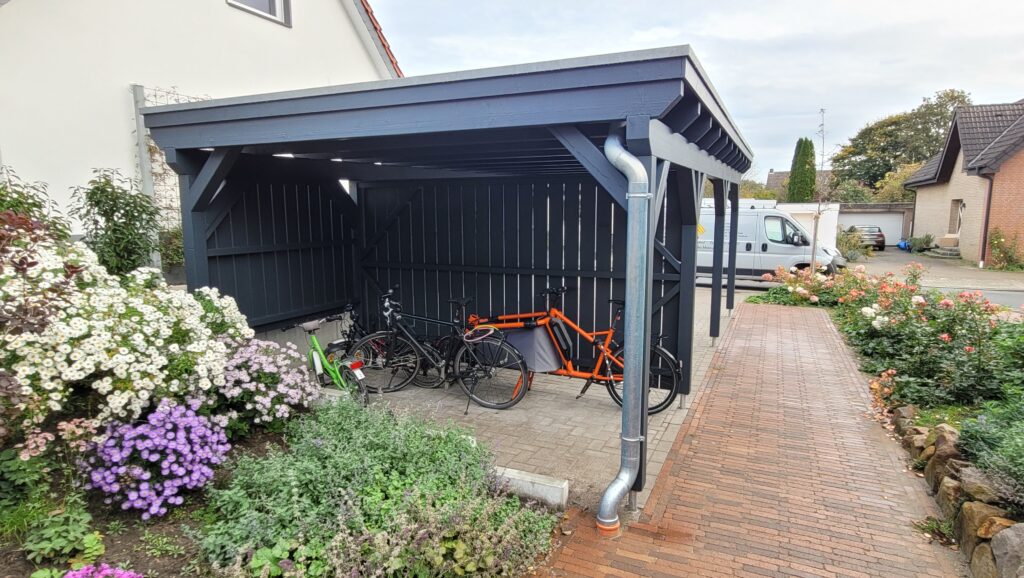 Ein überdachter Fahrradunterstand mit mehreren Fahrrädern, darunter ein oranges Lastenrad, neben Blumen.