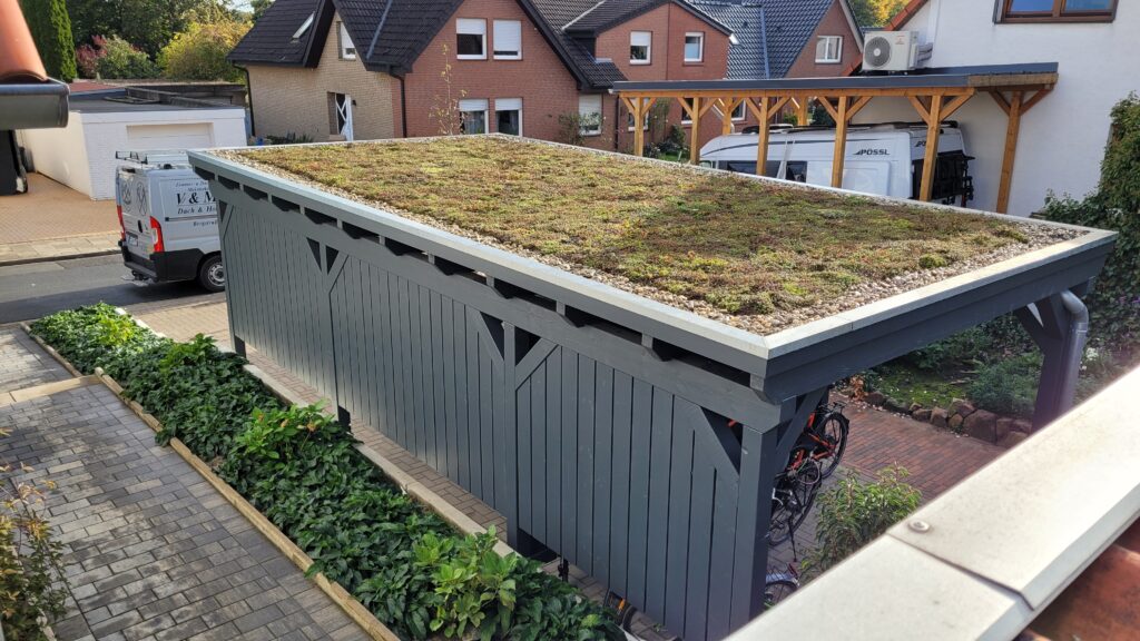 Ein modernes Carport mit begrüntem Dach, umgeben von Pflanzen und Wohnhäusern im Hintergrund.