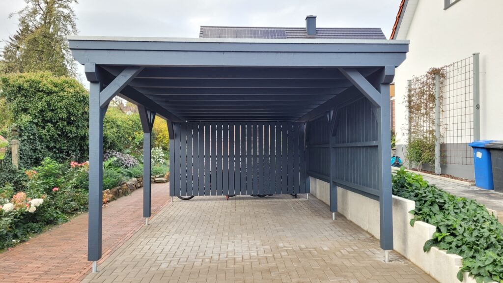 Moderner grauer Holz-Carport mit Solarpaneelen, umgeben von blühenden Pflanzen und gepflastertem Boden.