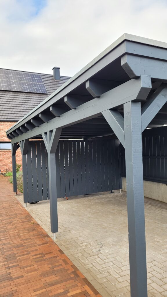 Moderner Carport aus dunklem Holz neben einem Ziegelgebäude mit Solarpanelen auf dem Dach.