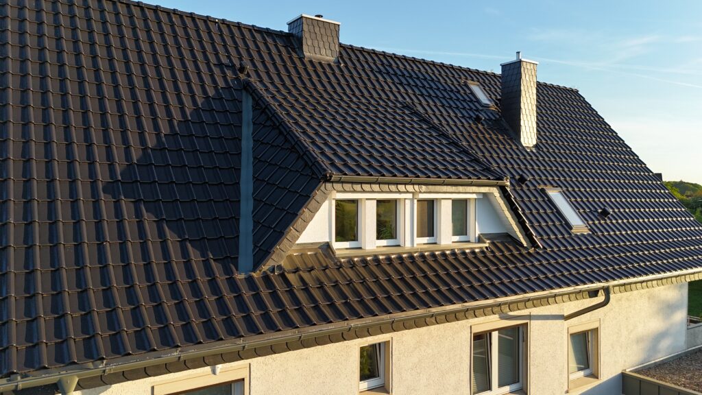 Ein Hausdach mit dunklen Ziegeln, zwei Schornsteinen und mehreren Dachfenstern unter klarem Himmel.