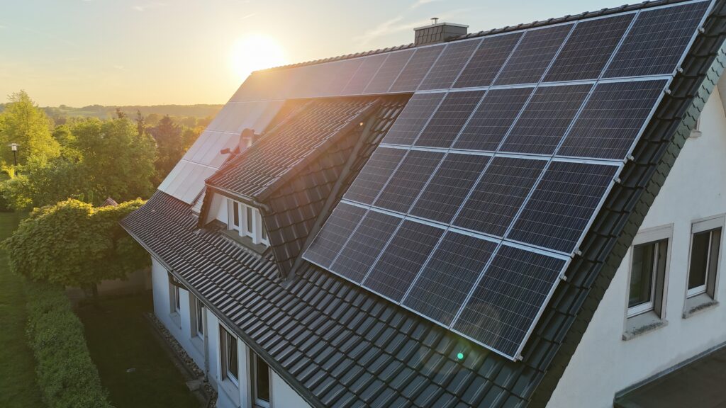 Ein Hausdach mit Solarpanelen bei Sonnenuntergang, umgeben von grüner Landschaft und klarem Himmel.