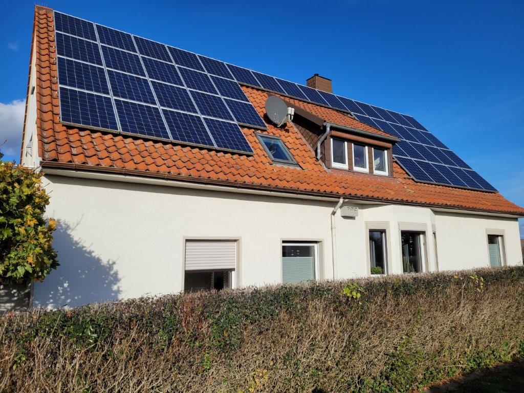 Ein Einfamilienhaus mit rotem Ziegeldach, bedeckt mit Solarpaneelen, umgeben von einer Hecke.