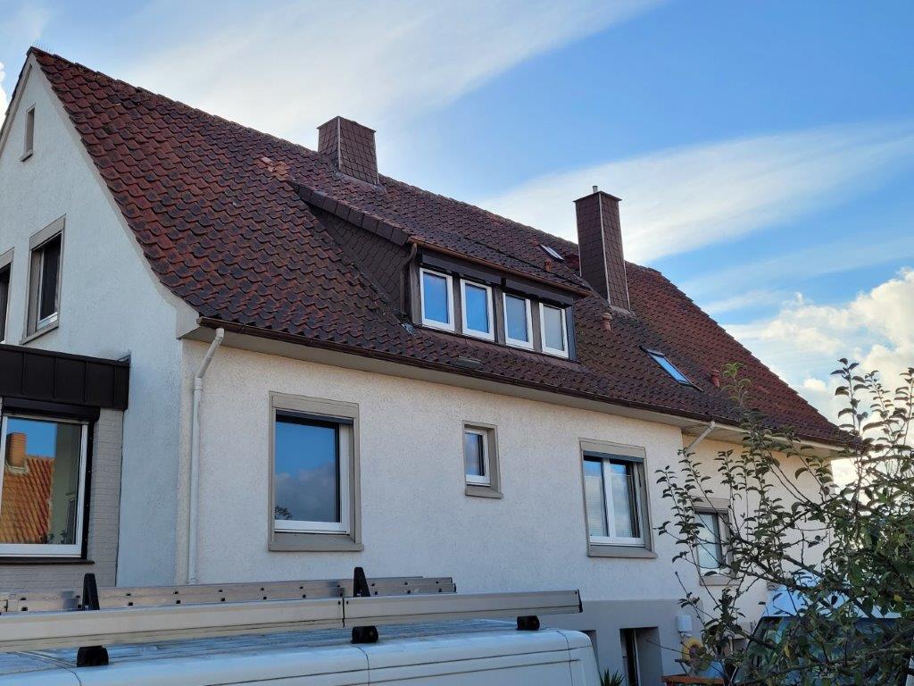 Seitenansicht eines Hauses mit rotem Ziegeldach, weißen Fenstern und einem Lieferwagen im Vordergrund.