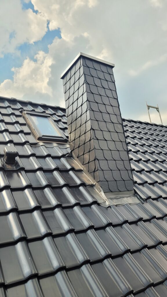 Ein Dach mit glänzenden, dunkelgrauen Ziegeln, einem schieferverkleideten Schornstein und einem Dachfenster.