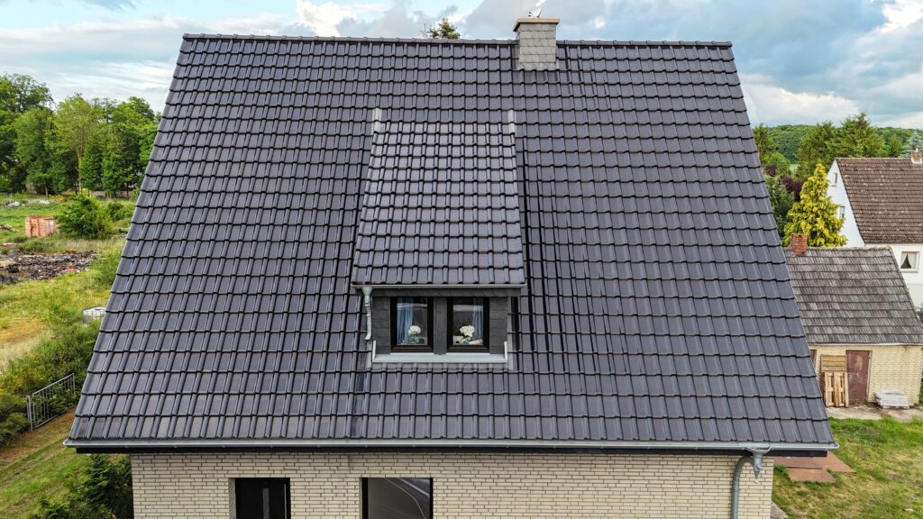 Ein Haus mit großem, dunkelgrauem Ziegeldach und Dachgaube, umgeben von Bäumen und Wiesen.