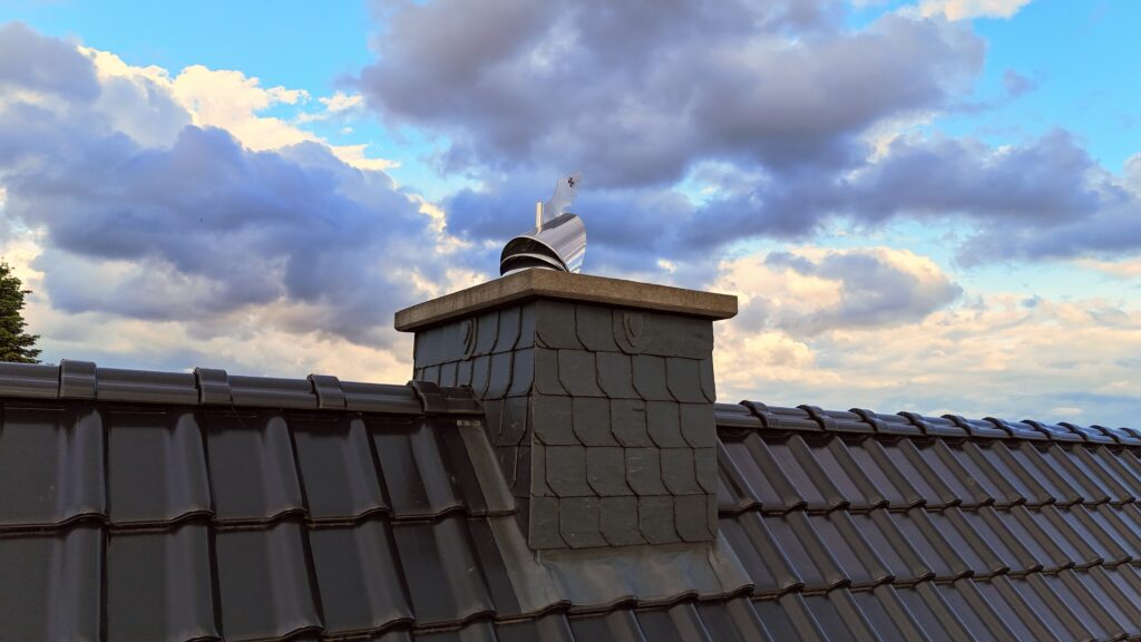 Ein Dach mit dunkelgrauen Ziegeln, einem Schornstein und einer silbernen Windturbine darunter bewölktem Himmel.
