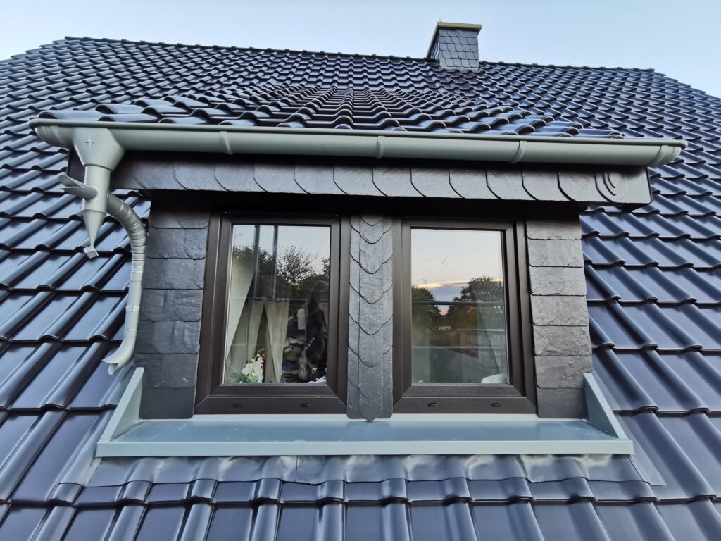 Ein Dach mit glänzenden dunkelblauen Ziegeln und einem zentralen Dachfenster mit dunklen Rahmen.