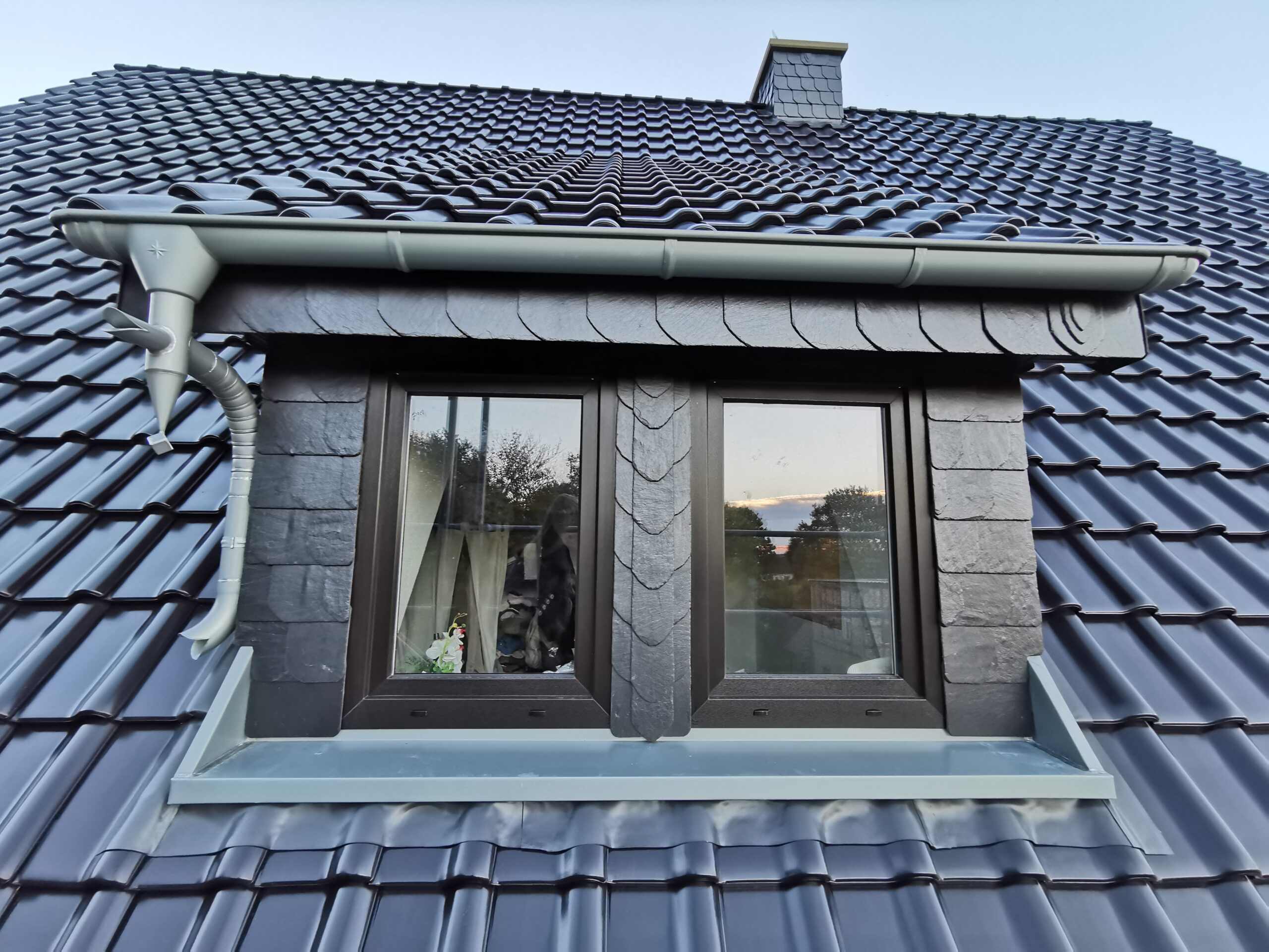 Ein Dach mit glänzenden dunkelblauen Ziegeln und einem zentralen Dachfenster mit dunklen Rahmen.