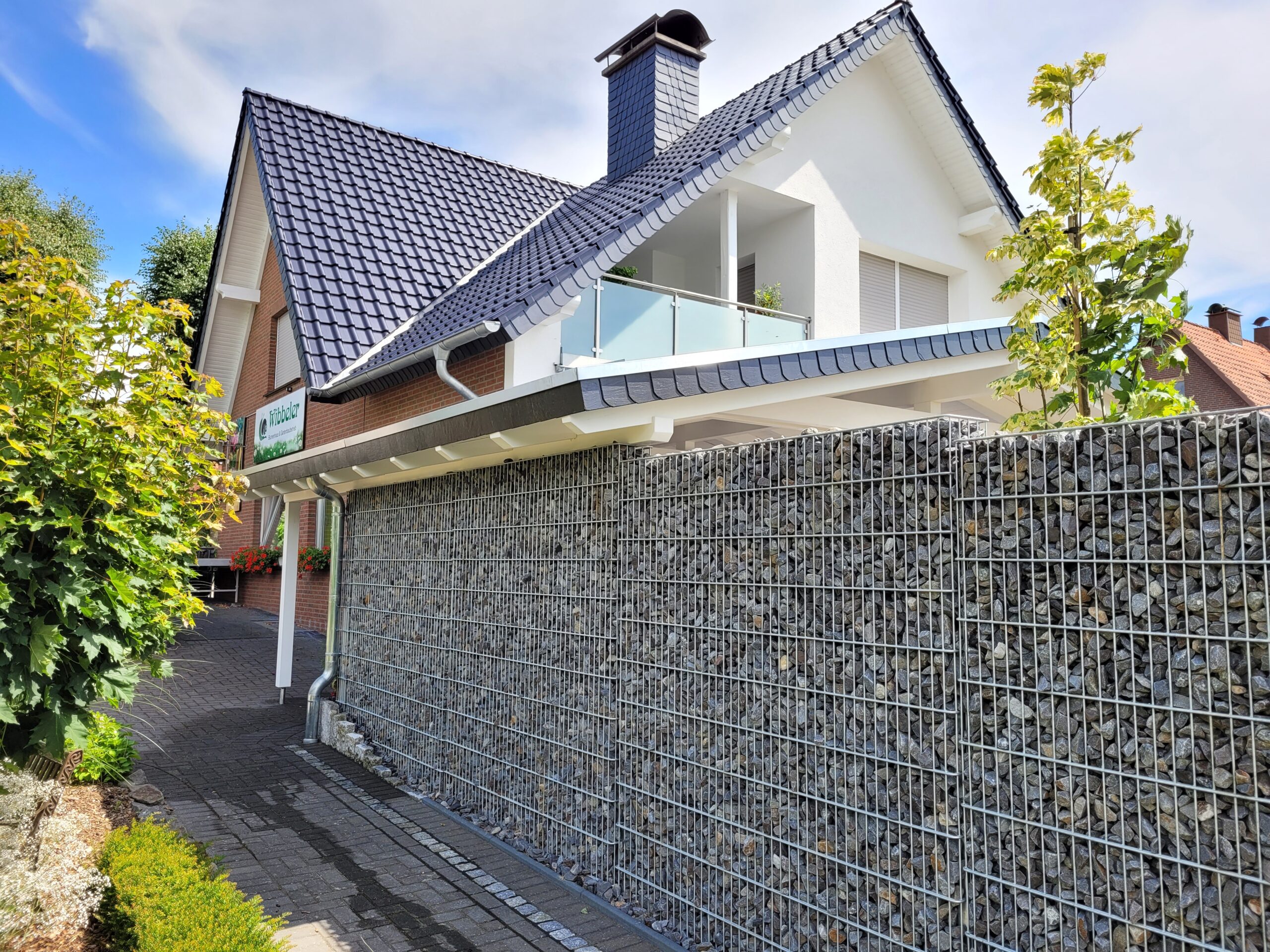 Modernes Wohnhaus mit steilem Dach, weißer Fassade, Glasbalkon und Gabionenwand, umgeben von Pflanzen.