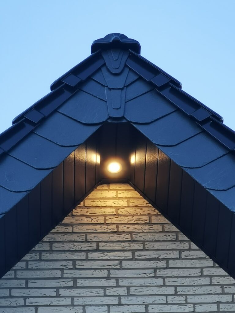 Giebel eines Hauses mit dunklem Dach, heller Ziegelwand und beleuchteter Außenlampe bei klarem Himmel.