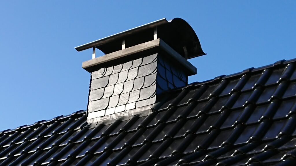 Ein Schornstein mit Schieferverkleidung auf einem Dach aus glänzenden, dunklen Ziegeln unter blauem Himmel.