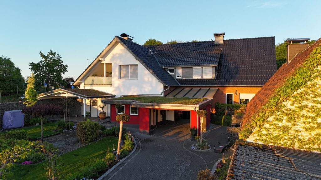Modernes Wohnhaus mit weiß-roter Fassade, Carport und gepflegtem Garten an einem sonnigen Tag.