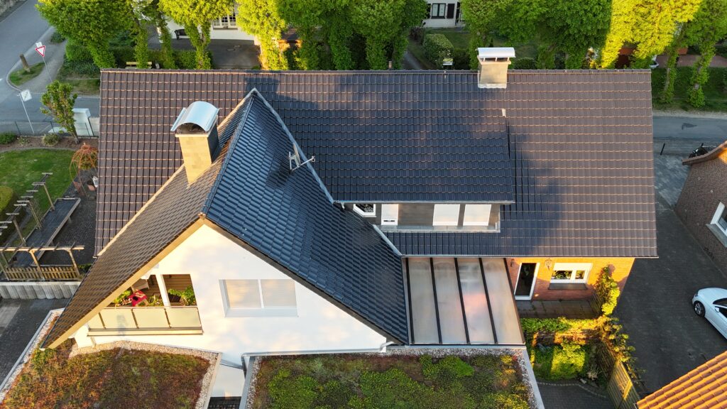 Luftaufnahme eines modernen Hauses mit dunklem Dach, Balkon, Carport und gepflegtem Garten.