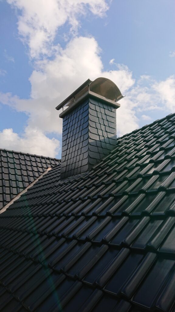 Ein Schornstein mit Schindeln auf einem dunklen Ziegeldach unter klarem, blauem Himmel mit Wolken.