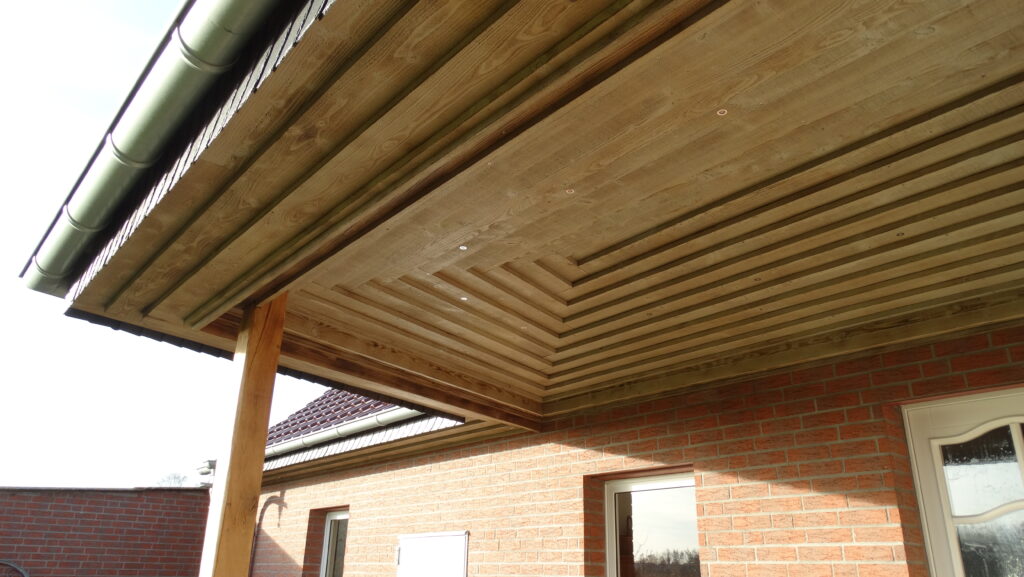 Überdachter Bereich mit Holzdecke, grüner Regenrinne und roter Ziegelwand mit Fenster und Tür.
