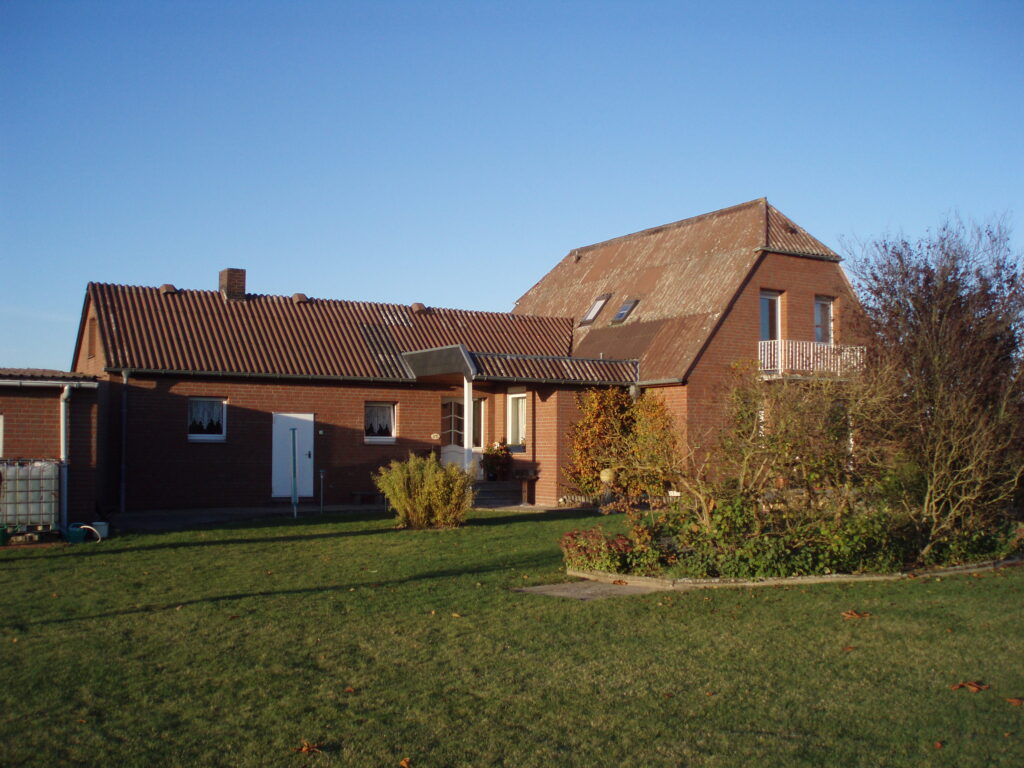 Ein rotes Ziegelhaus mit Balkon und Dachfenstern, umgeben von grünem Rasen und Sträuchern, unter blauem Himmel.