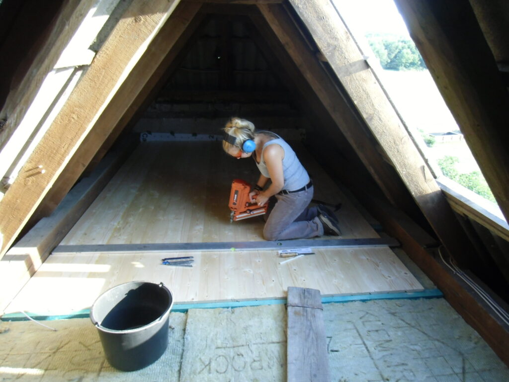 Eine Person arbeitet kniend auf einem Dachboden, befestigt Holzbretter mit einem Nagelgerät.