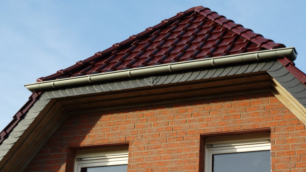 Ein Haus mit rotem Ziegeldach und Ziegelfassade, zwei Fenster unter dem Giebel, vor blauem Himmel.