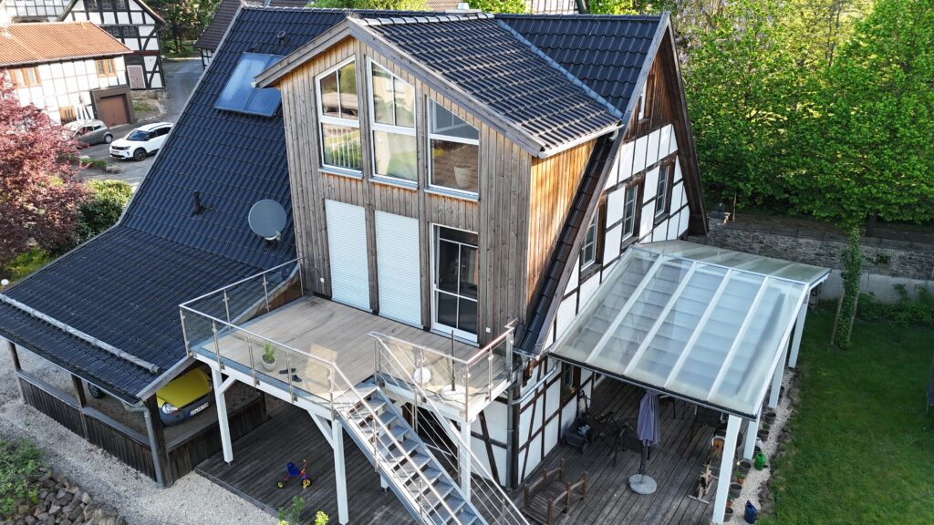 Modernes Fachwerkhaus mit Holz- und Glasfassade, großer Terrasse, Carport und umgeben von Natur.