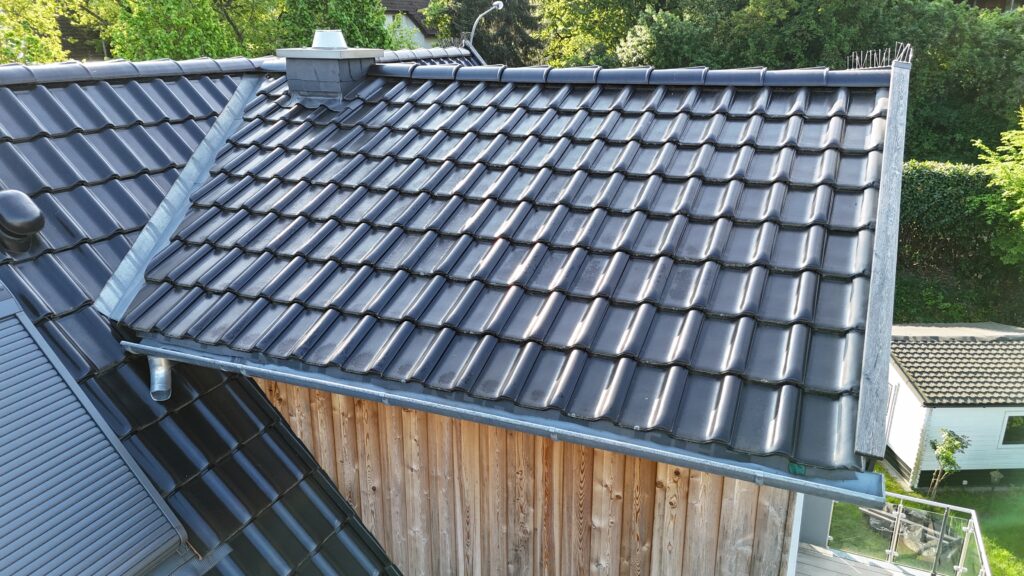 Dach mit glasierten, dunkelgrauen Ziegeln, kleinem Schornstein und Holzverkleidung, umgeben von Grün.