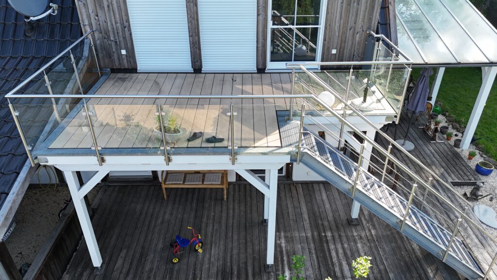 Moderne Terrasse mit Glasgeländer, Pflanzen, Treppe und Dreirad auf Holzboden, ruhige Atmosphäre.