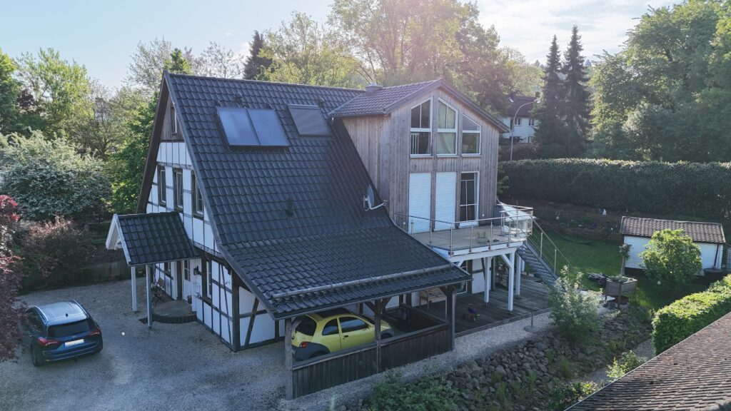 Modernes Fachwerkhaus mit Holz-Anbau, blauen und gelben Autos, Terrasse mit Glasgeländer, grüne Umgebung.