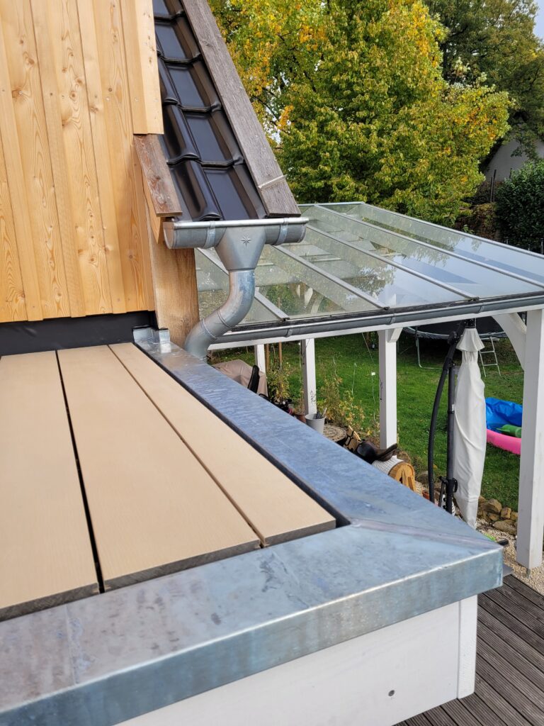 Ein Haus mit schwarzem Dach, Holzverkleidung, Terrasse und Glasdach, umgeben von Bäumen und Rasen.