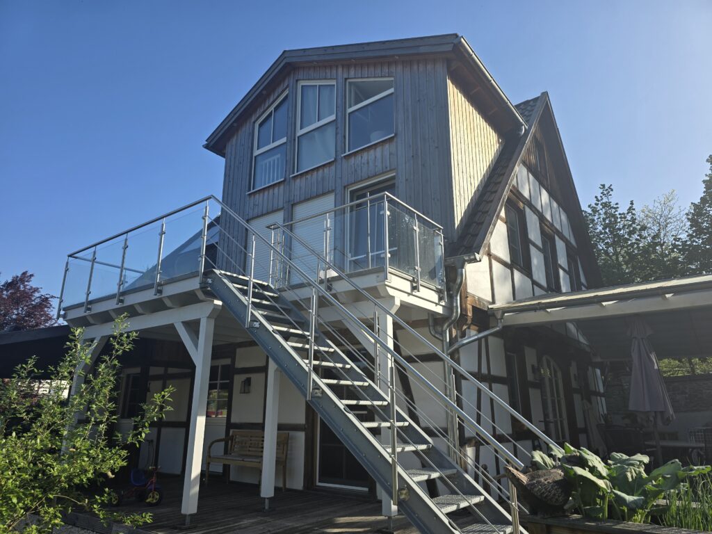 Modernes zweistöckiges Haus mit Fachwerk, Metalltreppe, Glasgeländer und Pflanzen im Vordergrund.