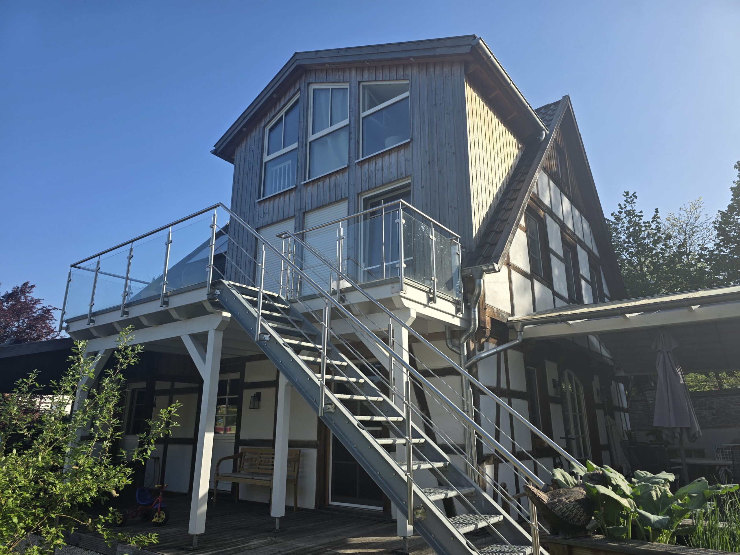 Modernes zweistöckiges Haus mit Fachwerk, Metalltreppe, Glasgeländer und Pflanzen im Vordergrund.