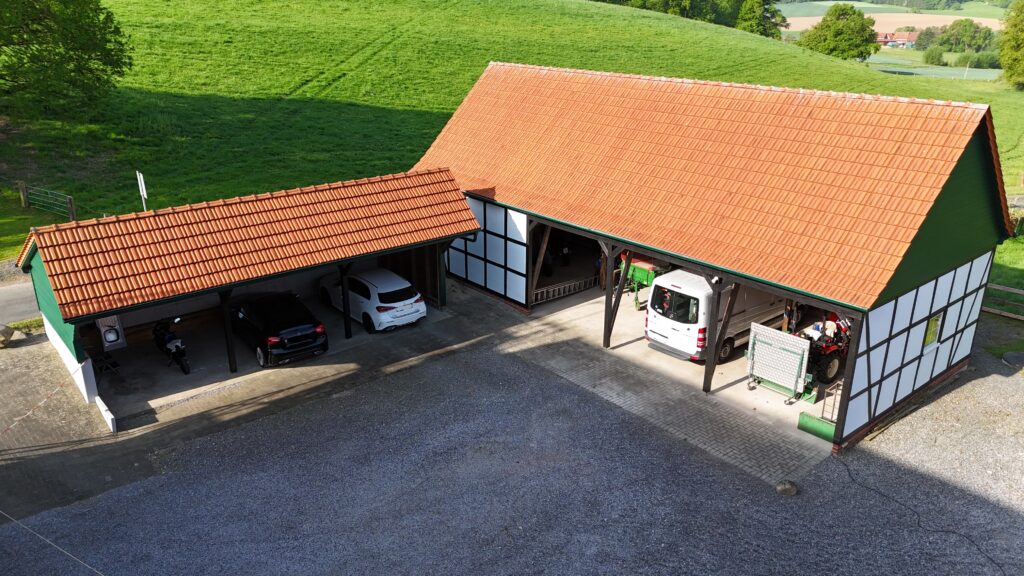 Ländlicher Hof mit Fachwerkgebäude, Carport und geparkten Fahrzeugen; ruhige, sonnige Atmosphäre.
