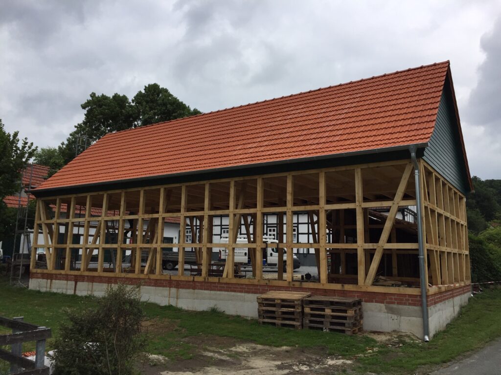 Unvollständiges Fachwerkgebäude mit Holzrahmen und rotem Ziegeldach, umgeben von Baumaterialien.