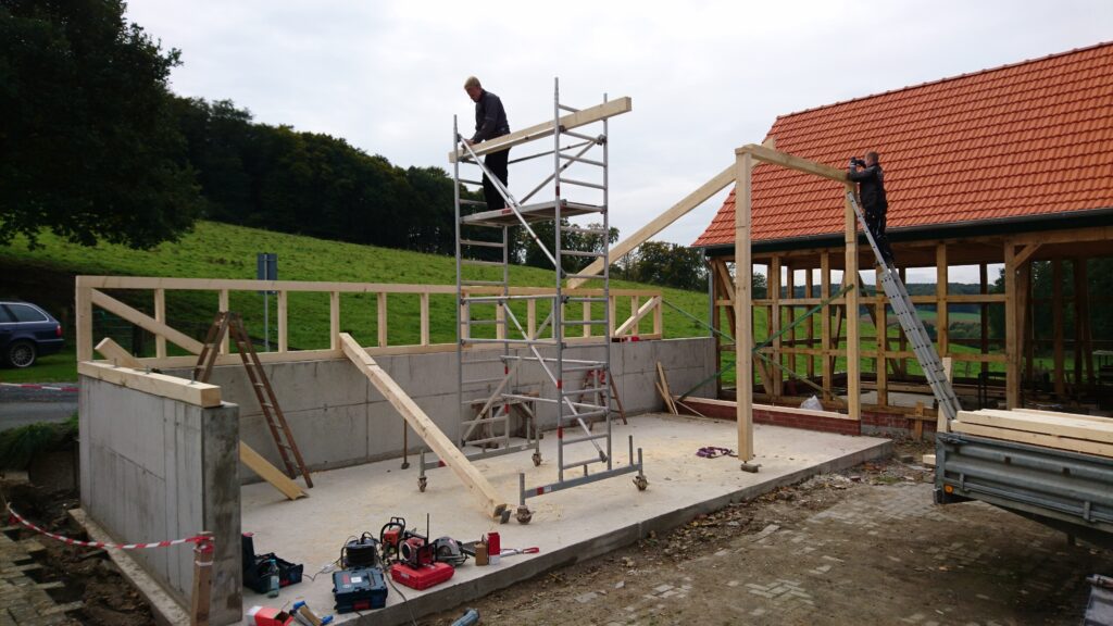 Zwei Personen arbeiten an einer Holzkonstruktion auf einer ländlichen Baustelle mit Ziegeldach im Hintergrund.