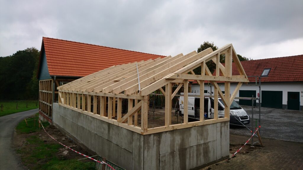 Baustelle mit Holzgerüst für Dach, bestehendes Gebäude mit rotem Ziegeldach im Hintergrund. Bewölkter Himmel.