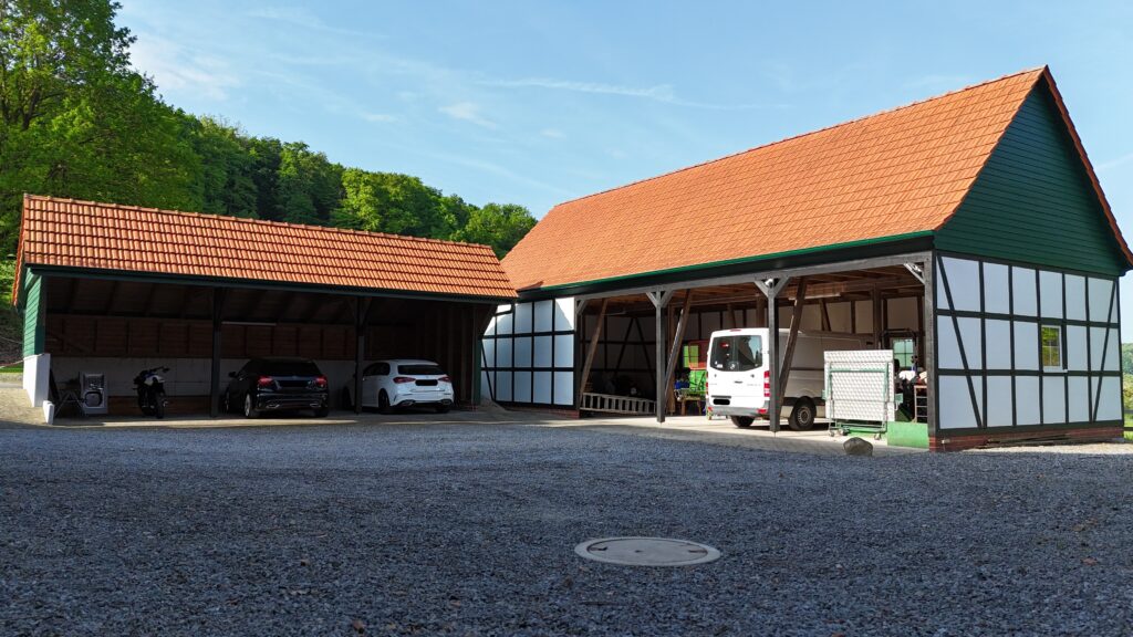 Ländlicher Hof mit Fachwerkgebäuden, roten Dächern, mehreren Fahrzeugen und klarem Himmel.
