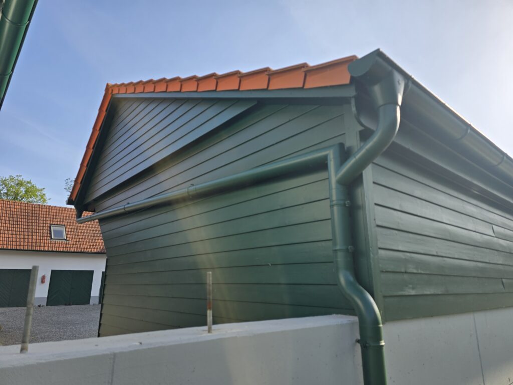 Grünes Gebäude mit rotem Ziegeldach, weiße Mauer im Vordergrund, blauer Himmel, sonniger Tag.